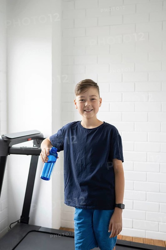 A young boy stands confidently by a treadmill, holding a blue water bottle. Dressed in casual sportswear, he smiles at the camera, ready for a workout in a bright room