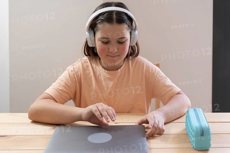 A young student with headphones on, concentrating on his laptop while learning online. The girl is preparing to start an online session, holding a pencil case on a wooden table