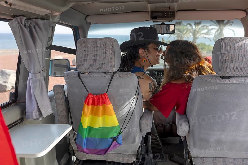 A lesbian couple enjoys a summer getaway in a van by the beach, with a rainbow pride bag in view. The scene captures freedom, love, and LGBTQIA+ pride