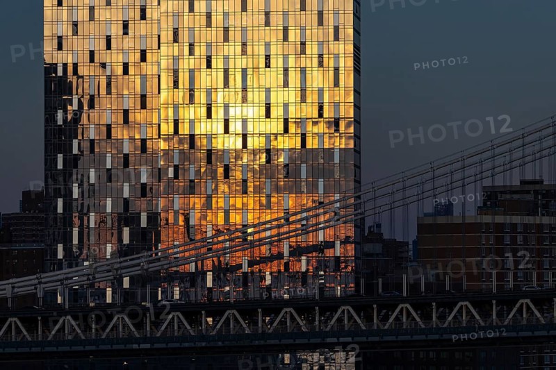 Golden sunlight reflects off a modern skyscraper in New York, creating a brilliant pattern contrasted against the darker silhouette of a nearby bridge