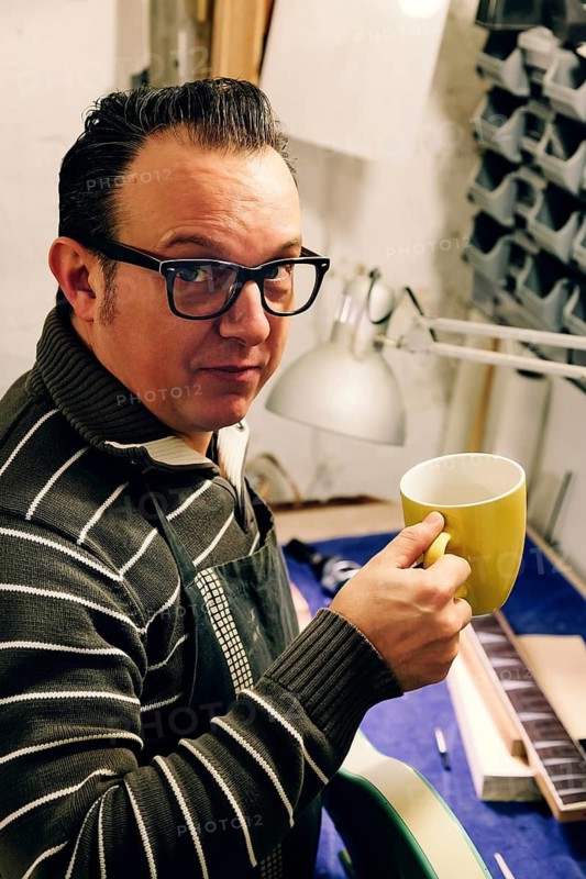 A skilled bass guitar luthier takes a coffee break in his cozy workshop. Surrounded by tools and instruments, he pauses from crafting a guitar neck for a moment of relaxation