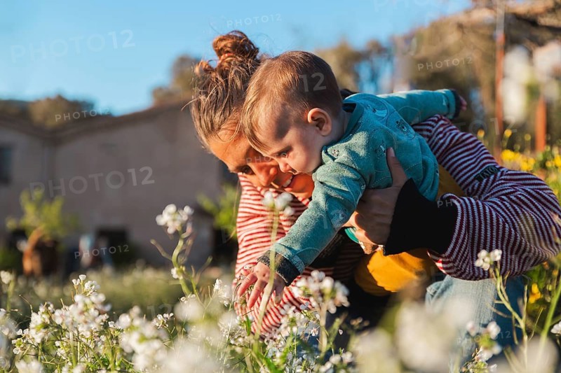A mother and her child enjoy a sunny day in a garden, exploring white flowers. The moment captures tender bonding and curiosity in a peaceful, natural environment