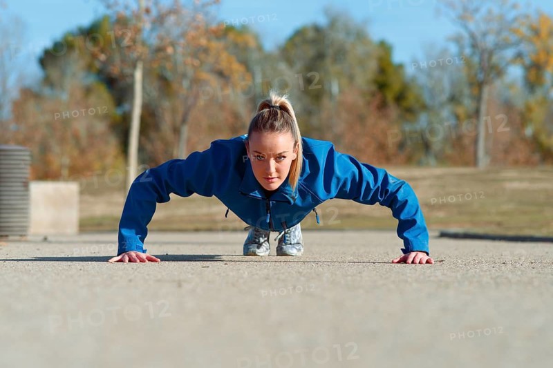 A woman in a blue jacket performs a push ups on a concrete surface outside, focused and determined. The background shows autumn trees under a clear blue sky