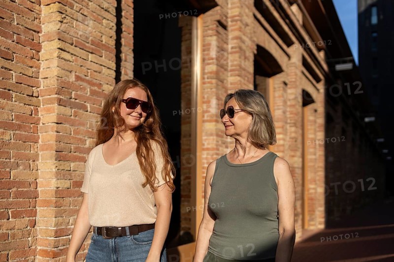 Mother and daughter walk together, enjoying a sunny day while sharing smiles and good company. The warm lighting and brick background create a heartfelt lifestyle moment