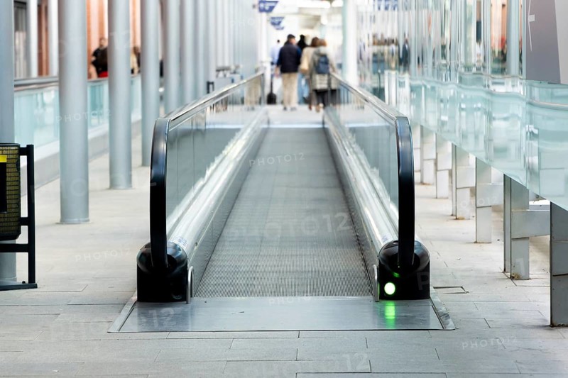 A contemporary urban scene featuring a moving walkway in a busy public transportation station with unrecognizable passengers in the background