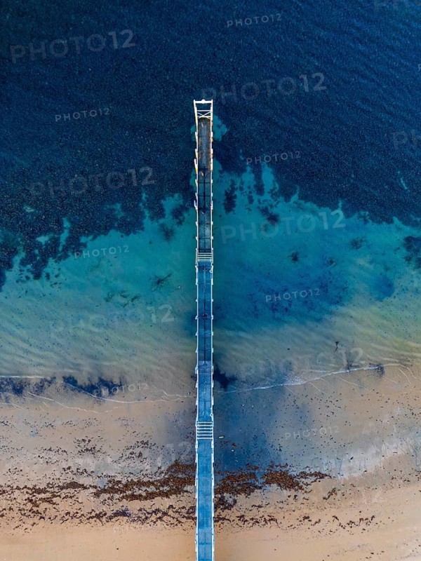A stunning aerial image shows a solitary pier extending into the vast, clear sea in the Great Ocean Road, Australia. The contrast between the deep blue water and sandy shore creates a serene scene
