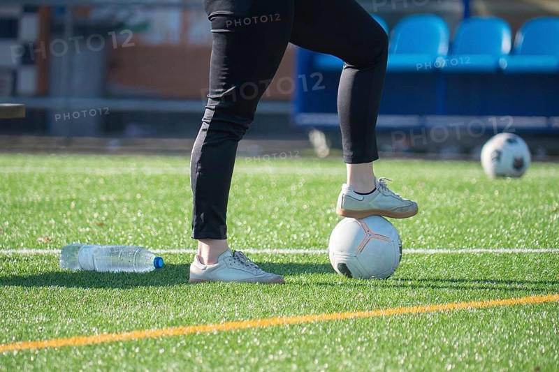A person wearing sneakers and black pants stands with one foot on a soccer ball on a grass soccer field. The scene includes another ball and a water bottle on the ground