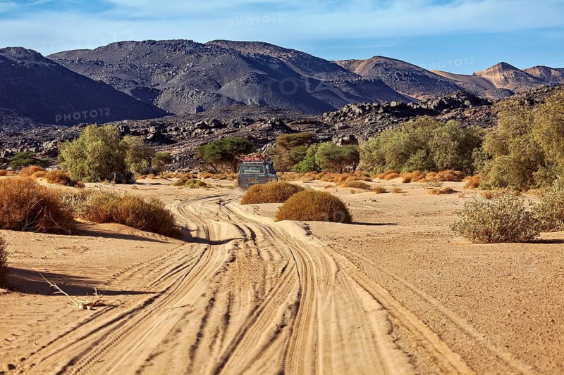 An arid desert landscape with a vehicle that leaves tracks in the sand and is surrounded by mountains, with an off-road vehicle through the landscape of the Sahara in Algeria