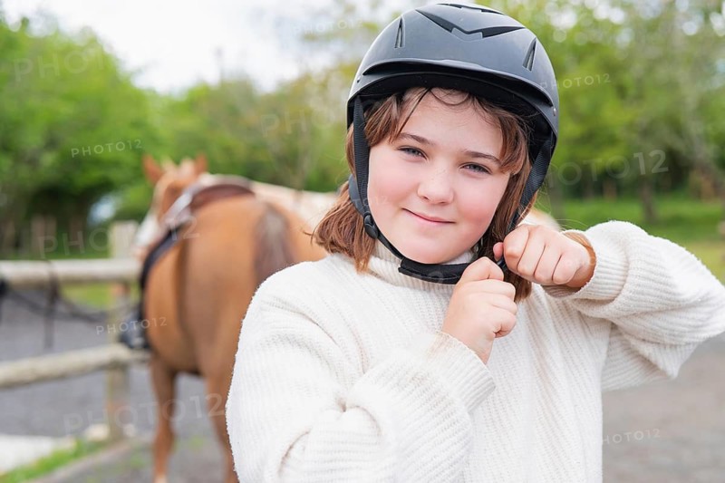 A young girl wearing a protective helmet fastens the strap, ready to enjoy a horse ride on a sunny day at the farm. Behind her, a brown horse stands calmly by the fence