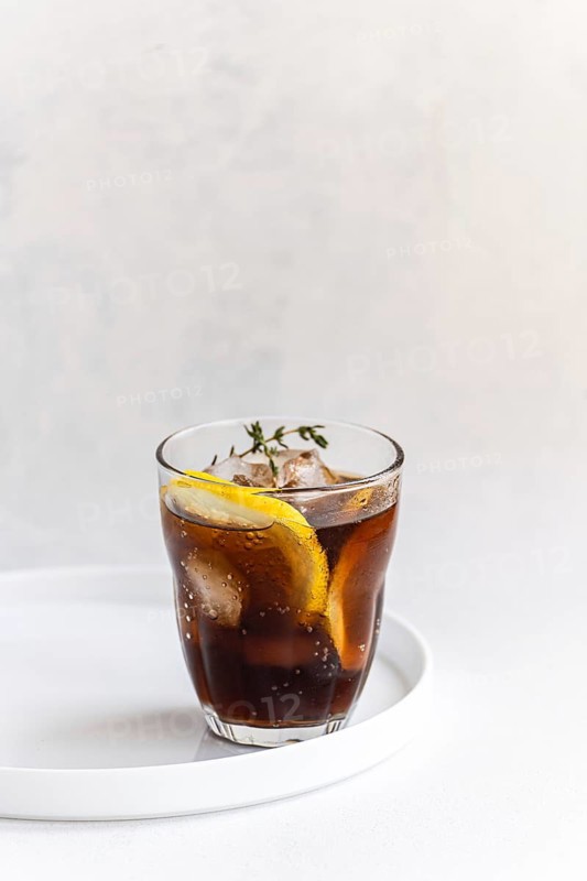A refreshing gin cocktail presented in a clear glass with cola drink, garnished with fresh thyme and a slice of lemon. Set against a simple white background, it exudes elegance and simplicity