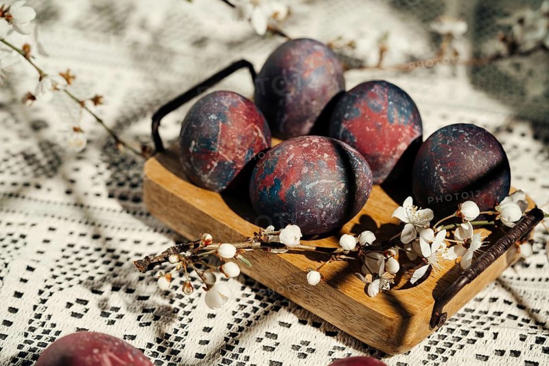 Artistic Easter eggs with a cosmic paint theme are displayed on a wooden tray surrounded by cherry blossoms, placed on a patterned crochet tablecloth, capturing a unique and festive springtime setting