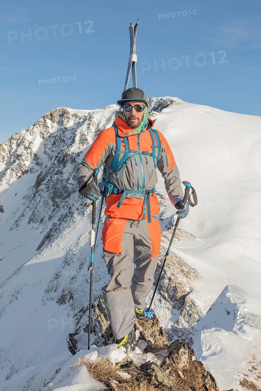 Ski tourer in bright gear standing on snowy ridge, basking in winter sunlight. The mountain view highlights the thrill of ski touring and adventure in alpine terrain