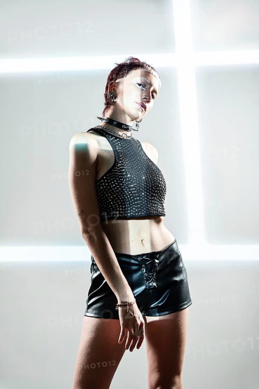 A striking cyberpunk woman model showcasing a futuristic look with a studded top and leather shorts, set against a glowing neon-lit background, embodying modern edginess