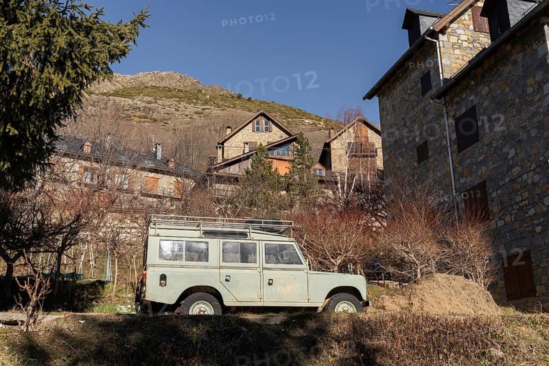 An old, classic off-road vehicle parked in front of traditional stone houses in a quaint, rural mountain village, surrounded by barren trees and hilly landscape