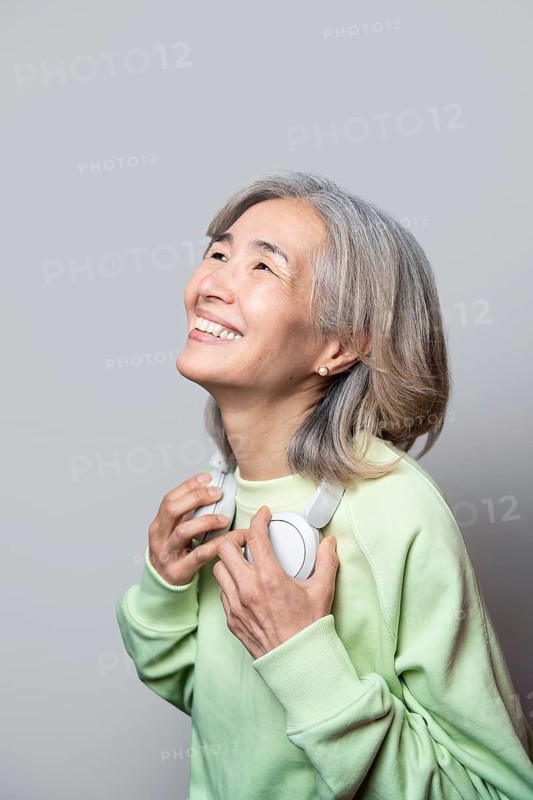 Asian senior woman wearing a light green sweater and headphones, looking up with a joyful expression. Her silver hair and serene smile convey a sense of peace and enjoyment