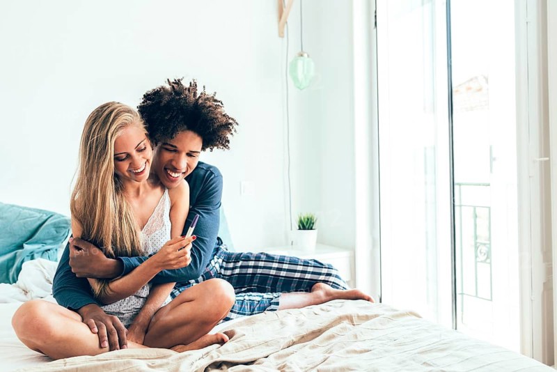 A happy couple sits on a bed, celebrating news while holding a pregnancy test. Warm light fills the bright room, capturing a moment of excitement and anticipation