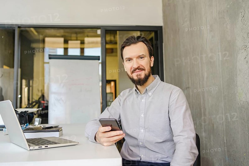 A man in a light shirt sits at a desk with a smartphone. A laptop and notepad accompany him in a bright, modern office environment