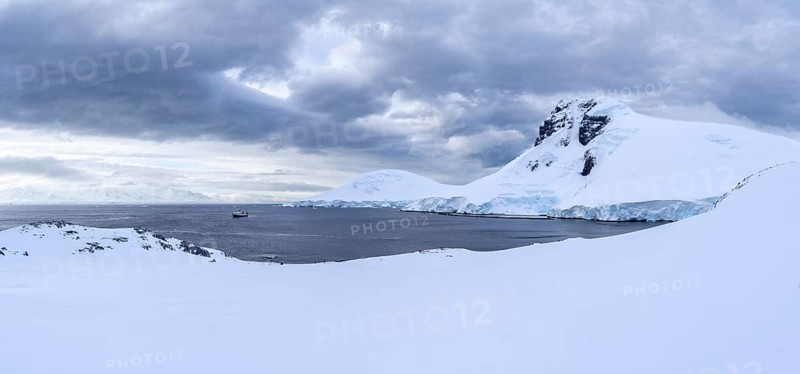 Snow-covered mountain landscape with glacier breakup on the coast, expedition ship anchored in the bay under cloudy sky, penguin colony in the foreground, Two Hummock Island, Palmer Archipelago, Antarctica