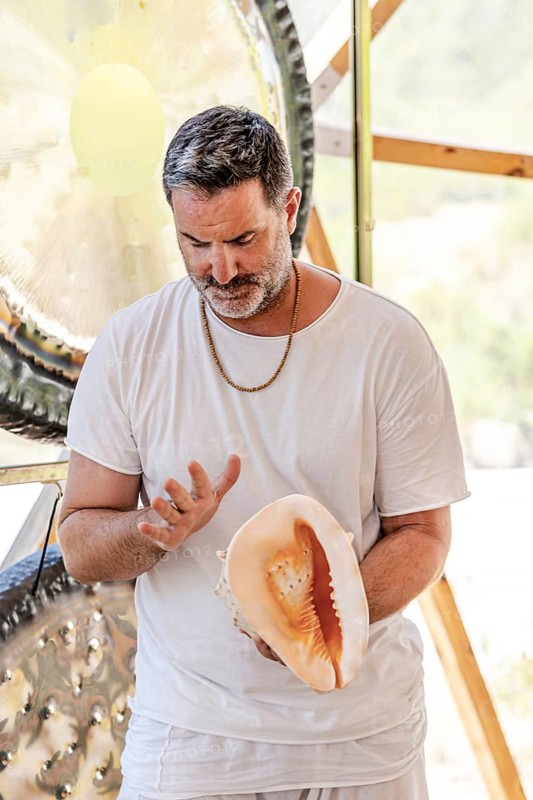 A male sound therapist practices sound therapy, using a conch shell in a serene setting. Observing traditional sound healing techniques for holistic wellness and relaxation