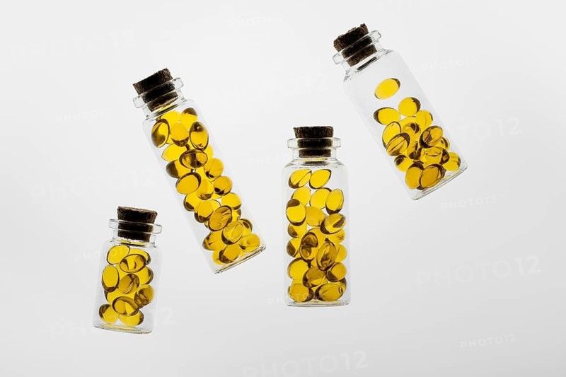 Transparent glass bottles filled with vibrant yellow Omega-3 and vitamin D capsules placed on a white background