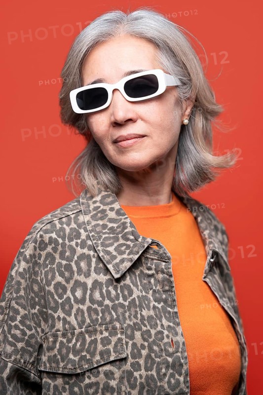 Asian senior woman poses confidently against a vibrant orange background. She wears trendy white sunglasses and a leopard print jacket, exuding modern fashion and flair