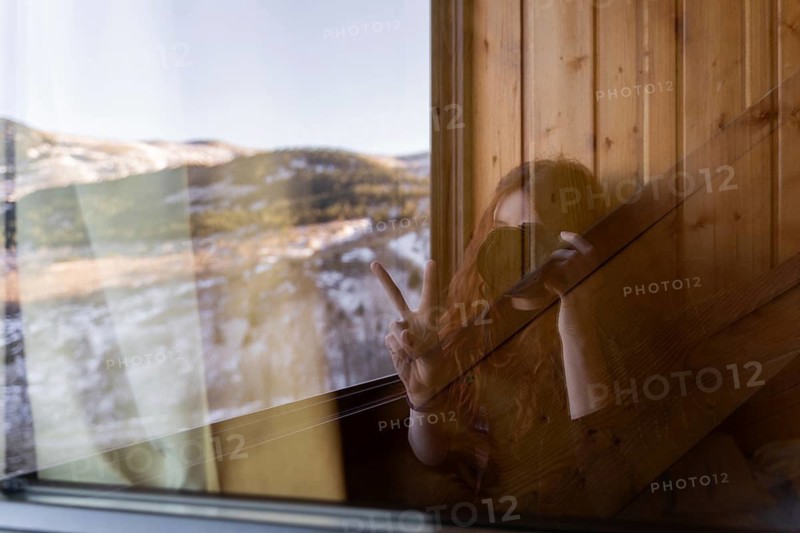 A redhead woman holding a camera is reflected in a cabin window, making a peace sign, with a snowy landscape visible in the background