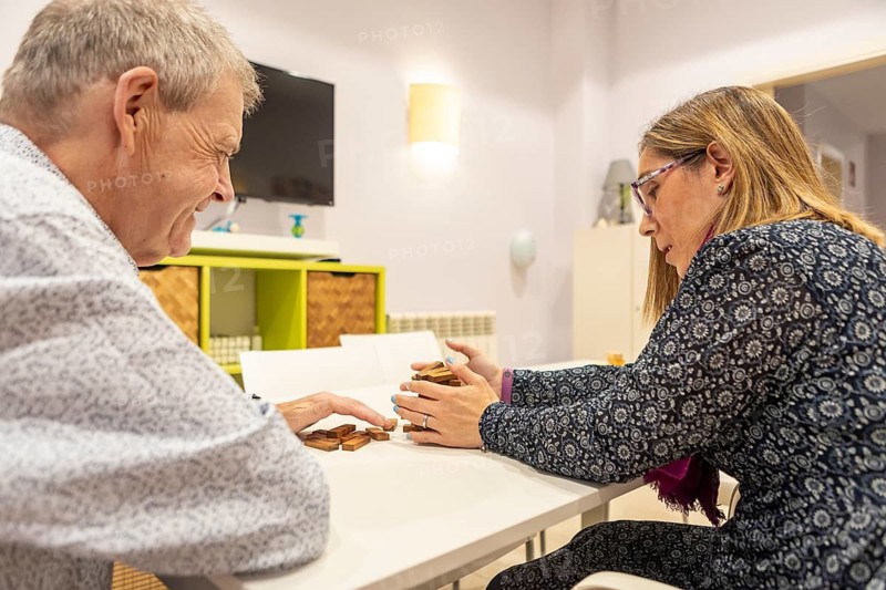 Couple with intellectual disabilities enjoy a board game at a table of a supervised home. Their expressions show concentration and joy as they engage in this stimulating and fun activity