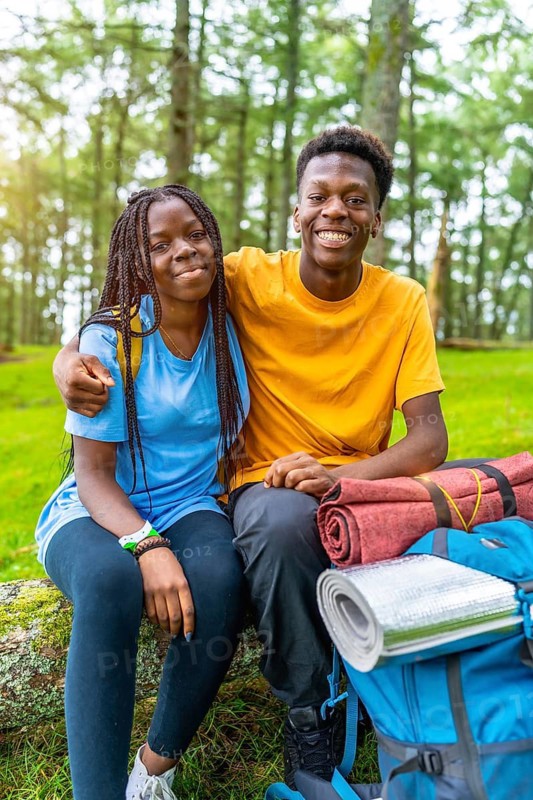 Vertical portrait of two young african friends sitting on fallen tree and resting during hiking