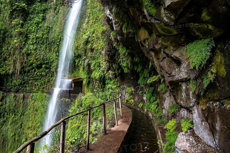 Path along the levada in the gorge of the Ribeira da Janela, below a rocky outcrop with wild vegetation and a waterfall, adventurous, Madeira, Portugal