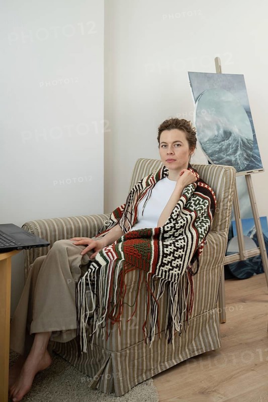A woman wearing a patterned shawl sits in a cozy armchair beside a wooden table with a laptop. A painted canvas rests on an easel in the bright room