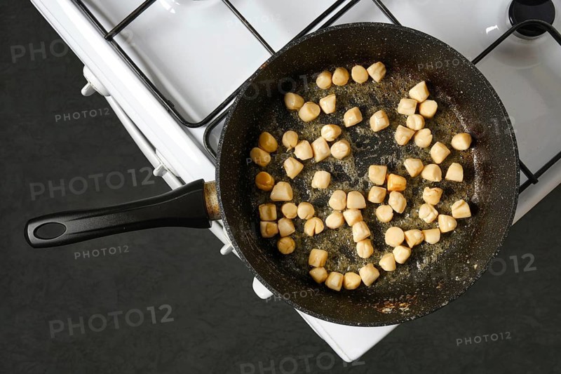 Scallops are being cooked in a frying pan on a stove. The pan is on a gas burner. The kitchen has a simple setup. Scallops are turning golden as they cook