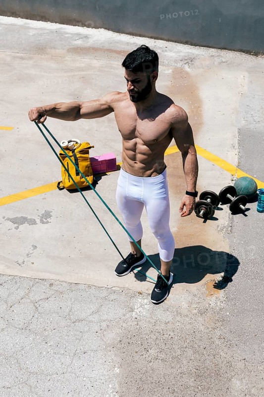 Fit man exercising with resistance bands outdoors, showcasing strength and athleticism. Dumbbells and sports equipment visible, emphasizing a healthy, active lifestyle