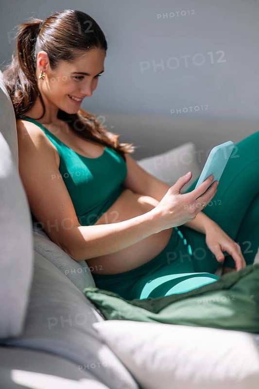 A pregnant woman in a comfortable yoga outfit sits on a couch, relaxed and focused on her smartphone. The setting exudes pregnancy wellness and serene mindfulness