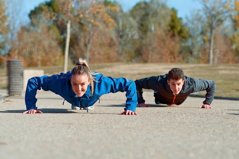 A man and woman doing push ups on a sunny day in a park. Both wear sportswear, showing determination and strength as they exercise on the pavement amid natural scenery