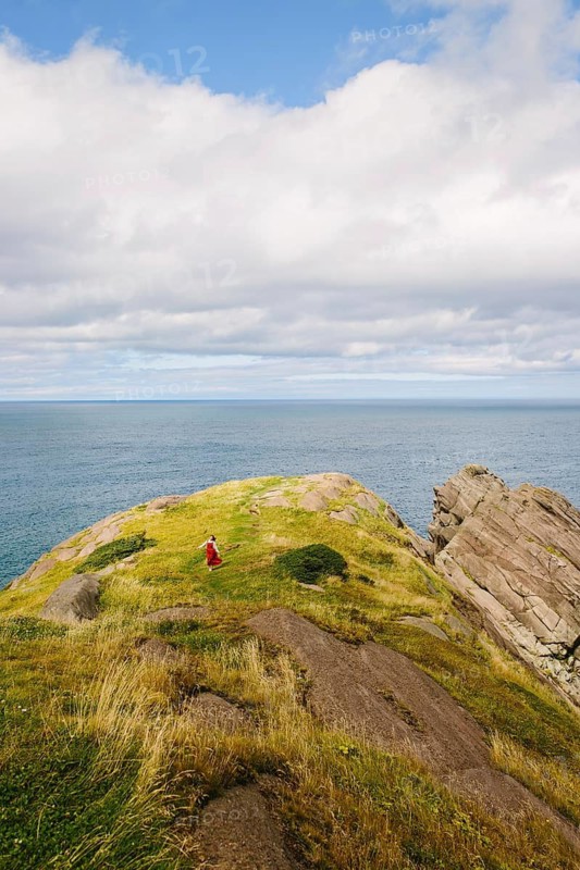 A female hiker in a vibrant red dress ascends a lush, rocky hillside at Signal Hill, Newfoundland & Labrador. The vastness of the ocean expands beyond under a partly cloudy sky, emphasizing the serene solitude and natural beauty of the scene