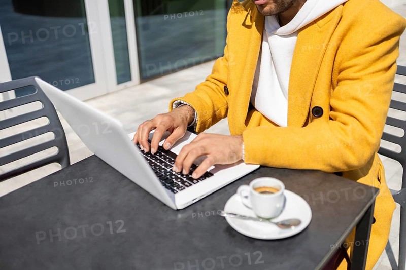 Afro man in a bright yellow coat types on a laptop at an outdoor cafe table on a sunny day, next to a small espresso cup, conveying a relaxed business setting