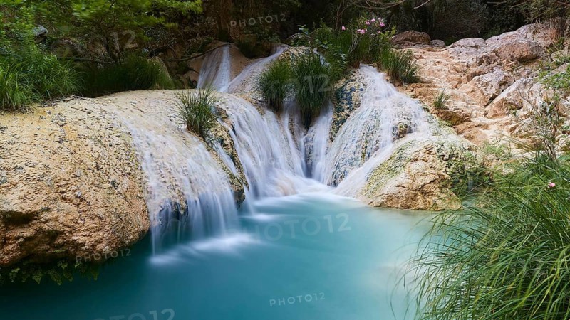 A small waterfall flows over rocks into a clear turquoise-blue pool surrounded by plants, waterfalls, ponds, Polylimnio, Peloponnese, Greece