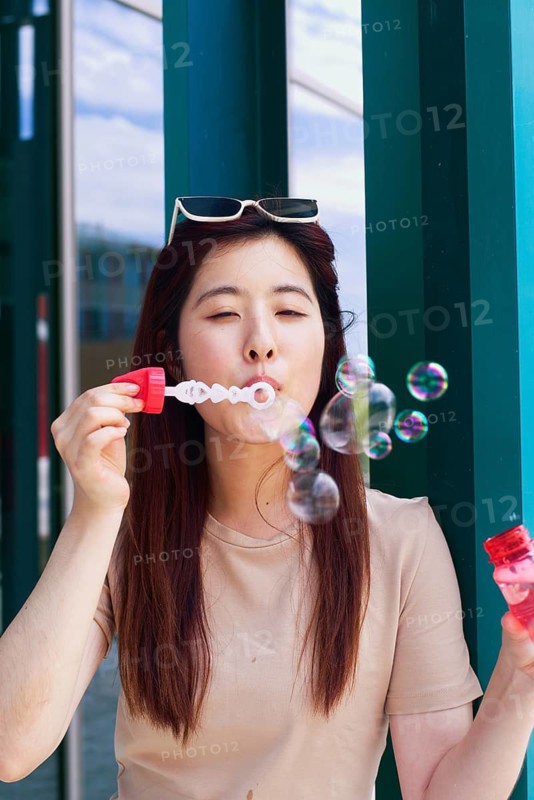 A young woman joyfully blows bubbles outside, capturing a playful moment. She wears sunglasses and a casual outfit, surrounded by colorful bubbles in the sunlight
