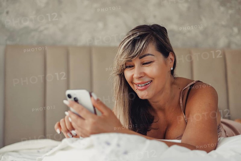 A smiling woman enjoys browsing her smartphone while lying comfortably on a bed. Her relaxed demeanor and engagement with the device create a warm and connected atmosphere