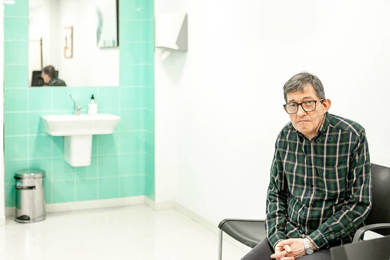 Man with glasses sits in a mental health facility, waiting for his appointment. The clinical setting with a sink and mirror reflects a calm, supportive environment focused on healthcare and well-being