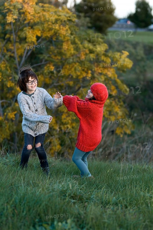In a lush park during Autumn, two siblings, a girl and a boy, enjoy playful moments surrounded by vibrant foliage. The girl in a red knitted sweater and a boy in gray play tag with joyful expressions