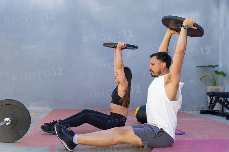 Two fitness professionals are performing a core strengthening exercise, lifting weights while sitting on mats in a gym's outdoor area, showcasing their dedication to physical wellness