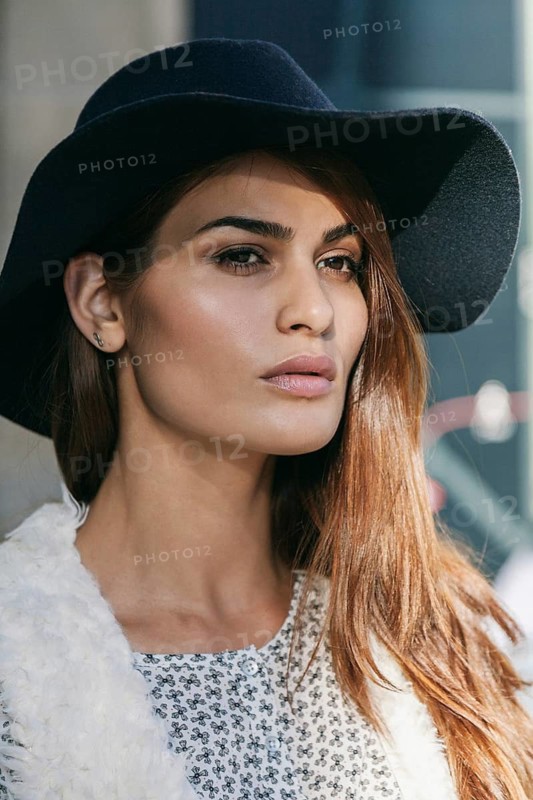 A woman wearing a wide-brimmed hat and a patterned blouse gazes thoughtfully. Her long hair falls naturally, highlighting her relaxed, stylish demeanor in natural light