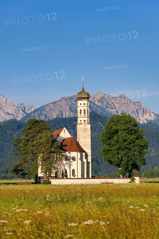 Church in the middle of a green meadow, surrounded by trees, with towering mountains in the background under a clear sky, Schwangau, Ostallgäu, Allgäu, Swabia, Upper Bavaria, Bavaria, Germany