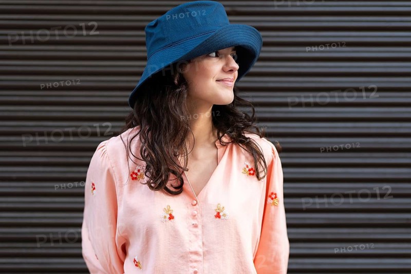 A young woman smiles confidently while wearing a stylish blue hat and a light pink blouse adorned with floral patterns. She stands against a striped outdoor background