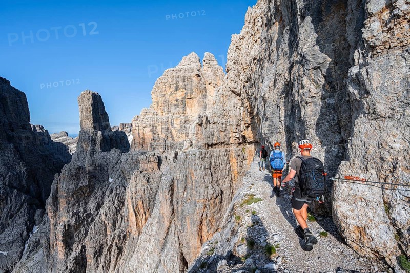 Three mountaineers on an exposed rock band in the secured Via Ferrata Bocciere Centrale via ferrata, spectacular mountain landscape with steep rocky peaks, Brenta Mountains, Parco Naturale Brenta-Adamello, Trentino, Italy