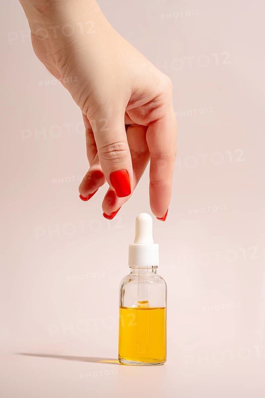 A hand with red manicured nails reaches for a clear glass dropper bottle filled with golden essential oil. The minimalistic beige background enhances the elegant aesthetic