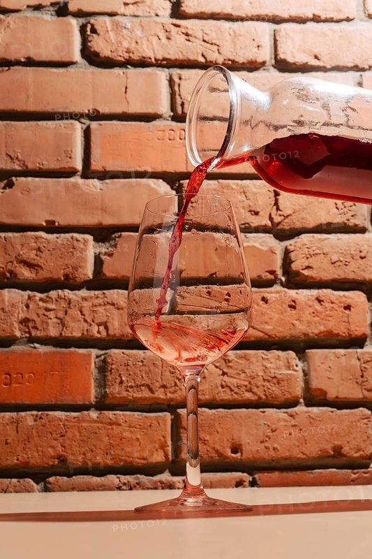 A stream of red wine flows from a glass carafe into a wine glass, set against a rustic brick wall. The warm tones highlight the rich color and texture of the wine