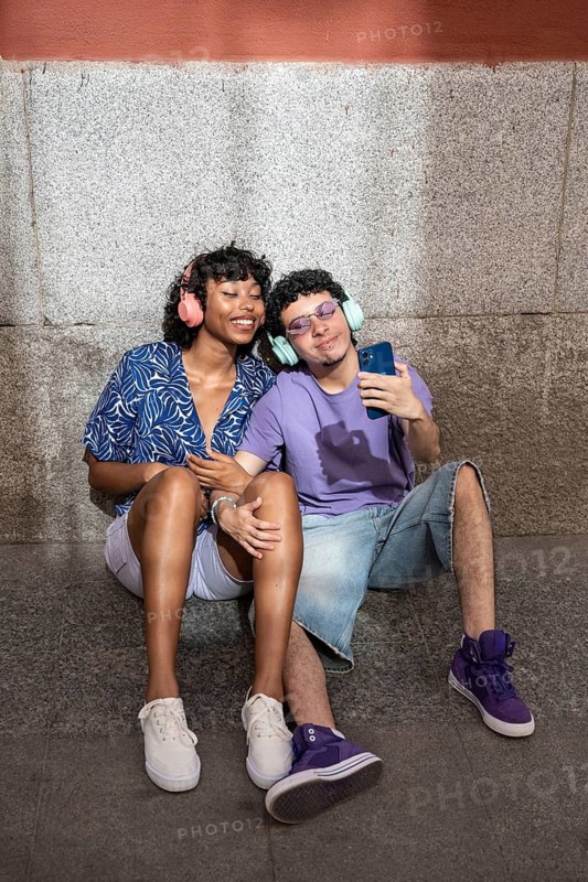 A joyful multiethnic LGBTQ+ couple sitting together, wearing headphones and sharing a smartphone, expressing connection, diversity, and love in an urban setting