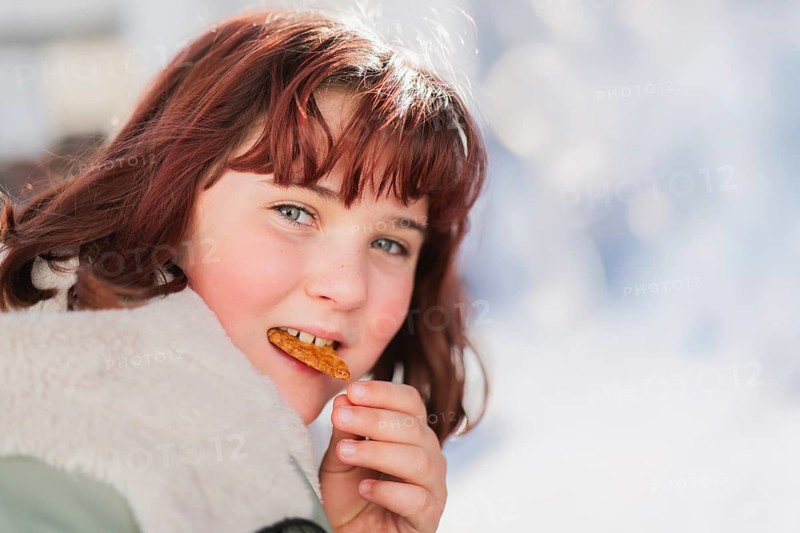 A young girl, with reddish-brown hair, enjoys a cookie on a bright winter day. Her warm coat and cheerful expression convey joy and warmth against the snowy backdrop